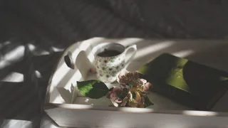 Plateau avec tasse de café et fleurs posé sur des draps gris, rayé par la lumière du matin