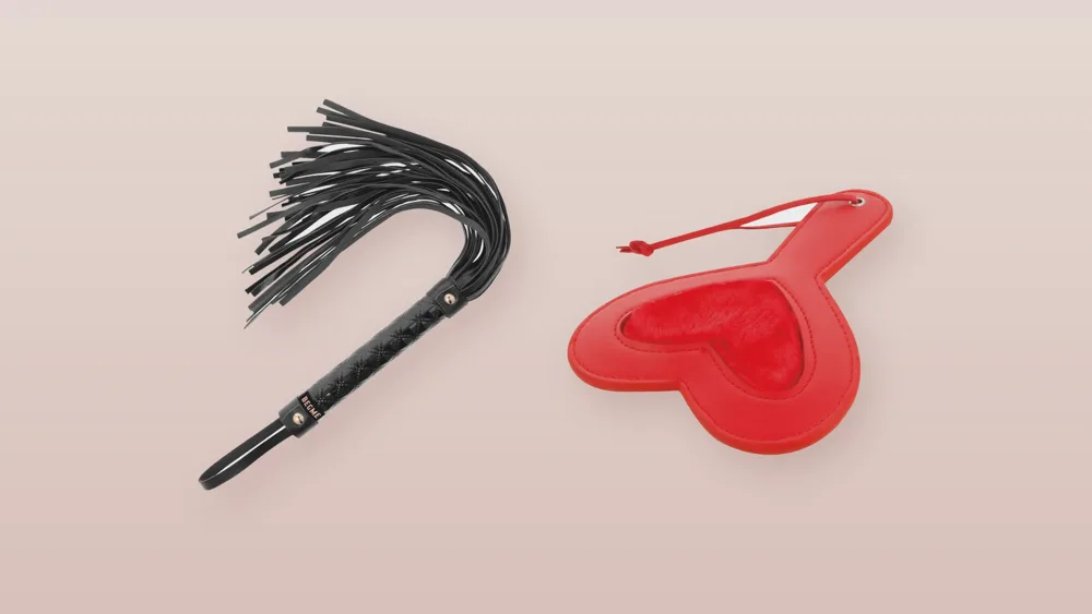 Martinet en simili-cuir noir et pagaie de fessée rouge en forme de cœur doublée de velours, posés sur un fond rose poudré