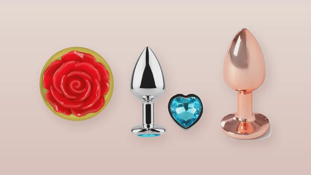 Trois bijoux anaux en métal sur fond rose poudré : base en rose rouge, strass bleu en forme de cœur et finition or rose