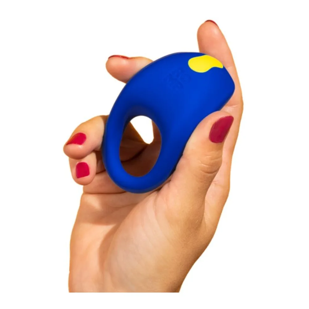 Anneau pénis en silicone bleu électrique avec détails jaunes et rouges, tenu en main avec des ongles vernis, présentant une forme ergonomique avec ouverture centrale