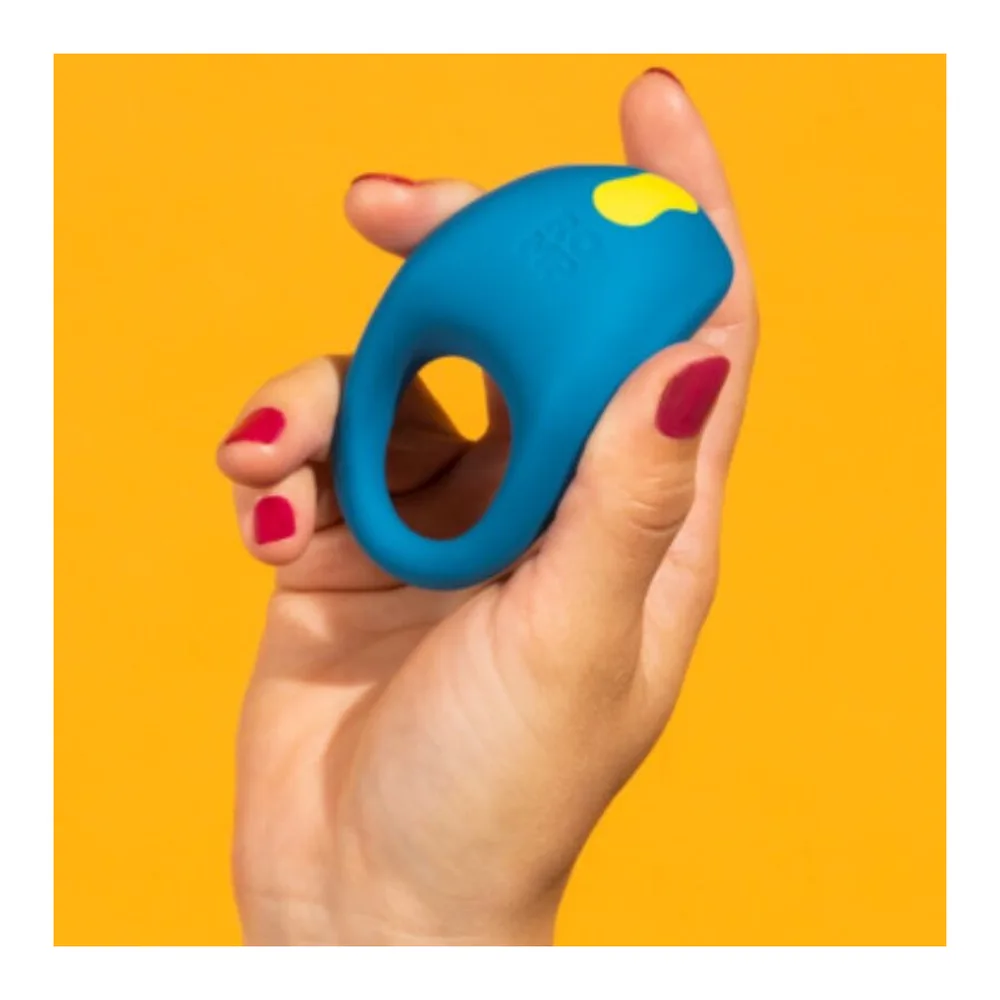 Anneau pénis en silicone bleu électrique avec détails jaunes et rouges, tenu en main avec des ongles vernis, présentant une forme ergonomique avec ouverture centrale