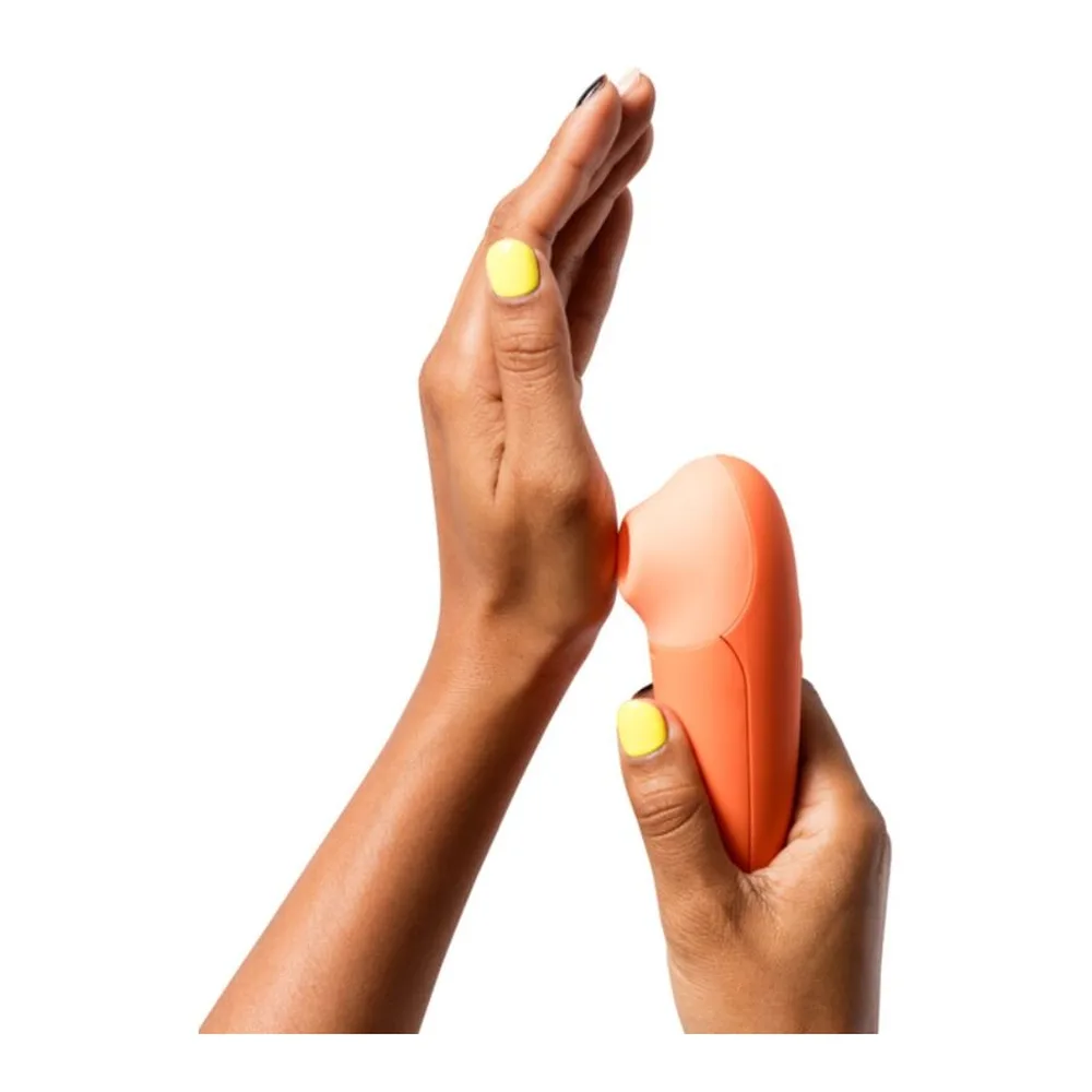 Stimulateur clitoridien orange porté à la main, tête flexible inclinée, avec un doigt aux ongles jaunes appuyant dessus, sur fond blanc