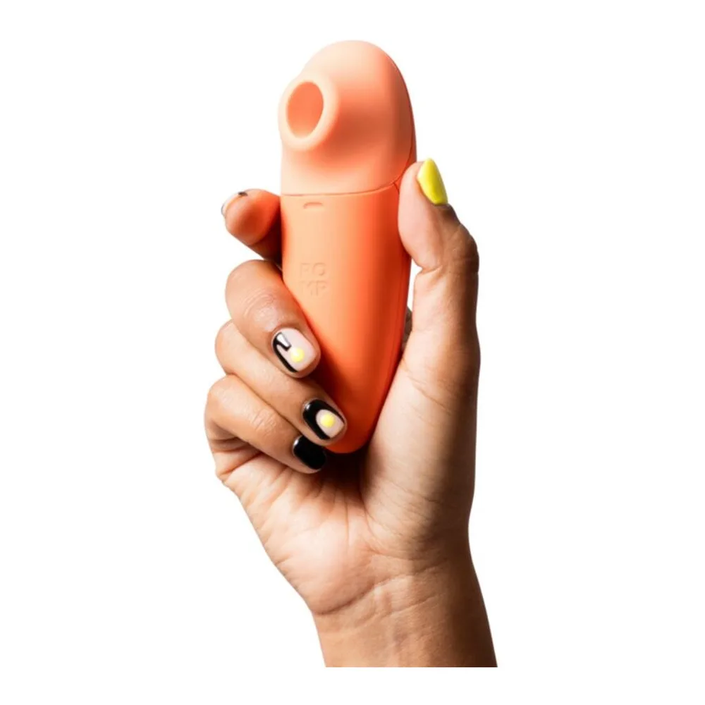 Stimulateur clitoridien orange porté à la main, tête flexible inclinée, avec un doigt aux ongles jaunes appuyant dessus, sur fond blanc