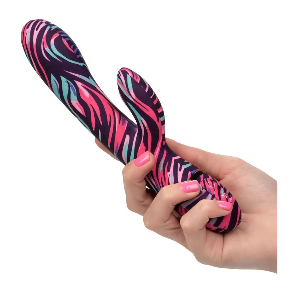 Vibromasseur rabbit en silicone rose, violet et bleu à motifs zébrés, avec deux branches flexibles, tenu en main sur fond blanc