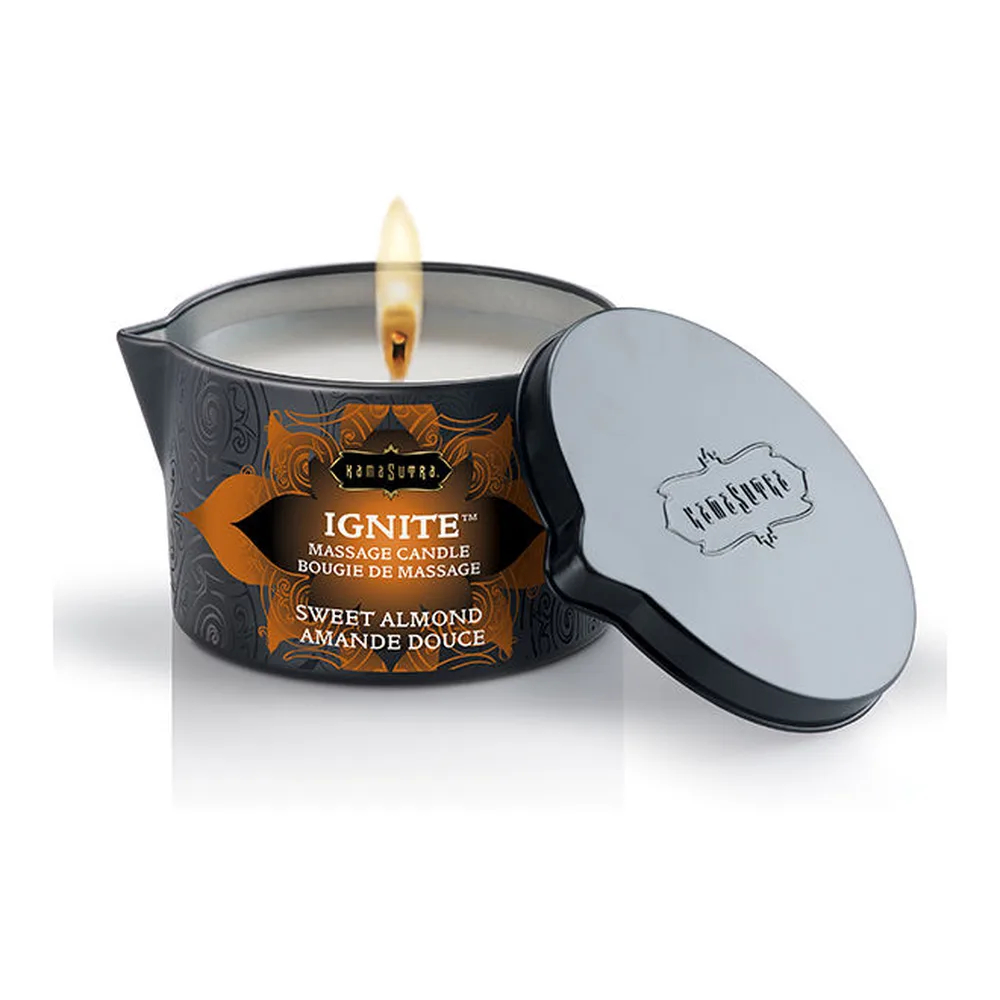 Bougie de massage Ignite en pot noir à motifs orangés, allumée avec flamme visible, accompagnée de son couvercle gris clair et cuillère. Étiquette imprimée indiquant Sweet Almond, sur fond blanc