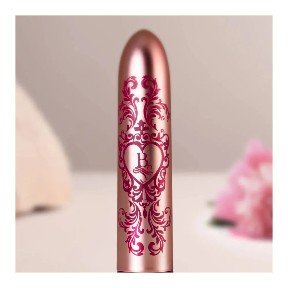 Stimulateur clitoridien de forme allongée en silicone couleur rose métallisé, orné de motifs floraux damassés en rose foncé, avec anneau de commande à la base, sur fond blanc