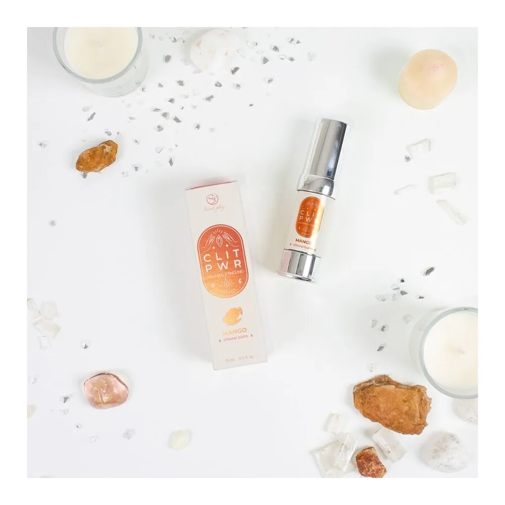 Baume clitoridien Clit Pwr en tube blanc et flacon applicateur à bille, entouré de cristaux d'ambre, de lait et de mangue sur fond blanc