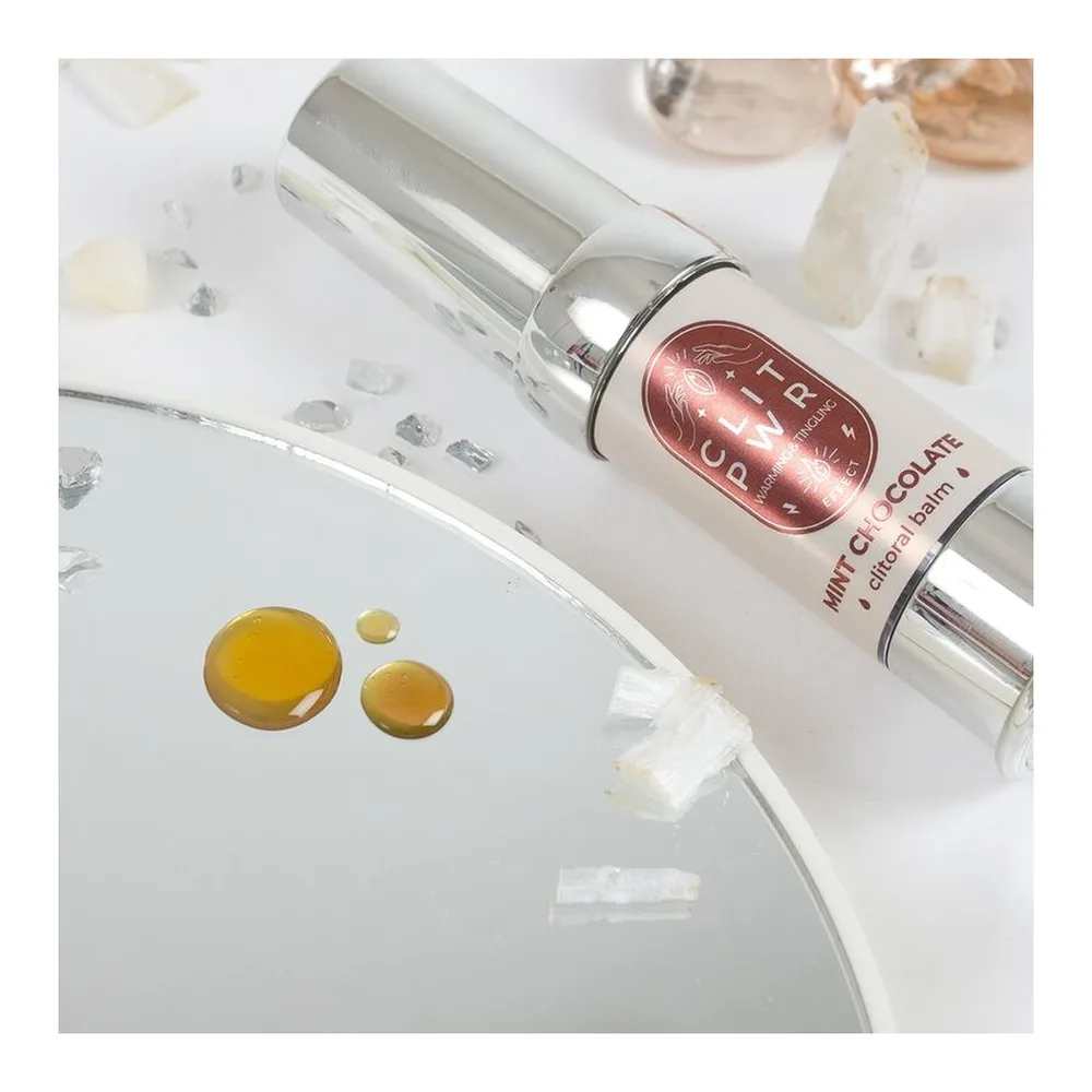 Baume clitoridien chauffant Clit Pwr parfum chocolat-menthe : emballage cartonné beige avec étiquette bordeaux, et tube applicateur blanc à pompe chromatée tenu en main sur fond blanc