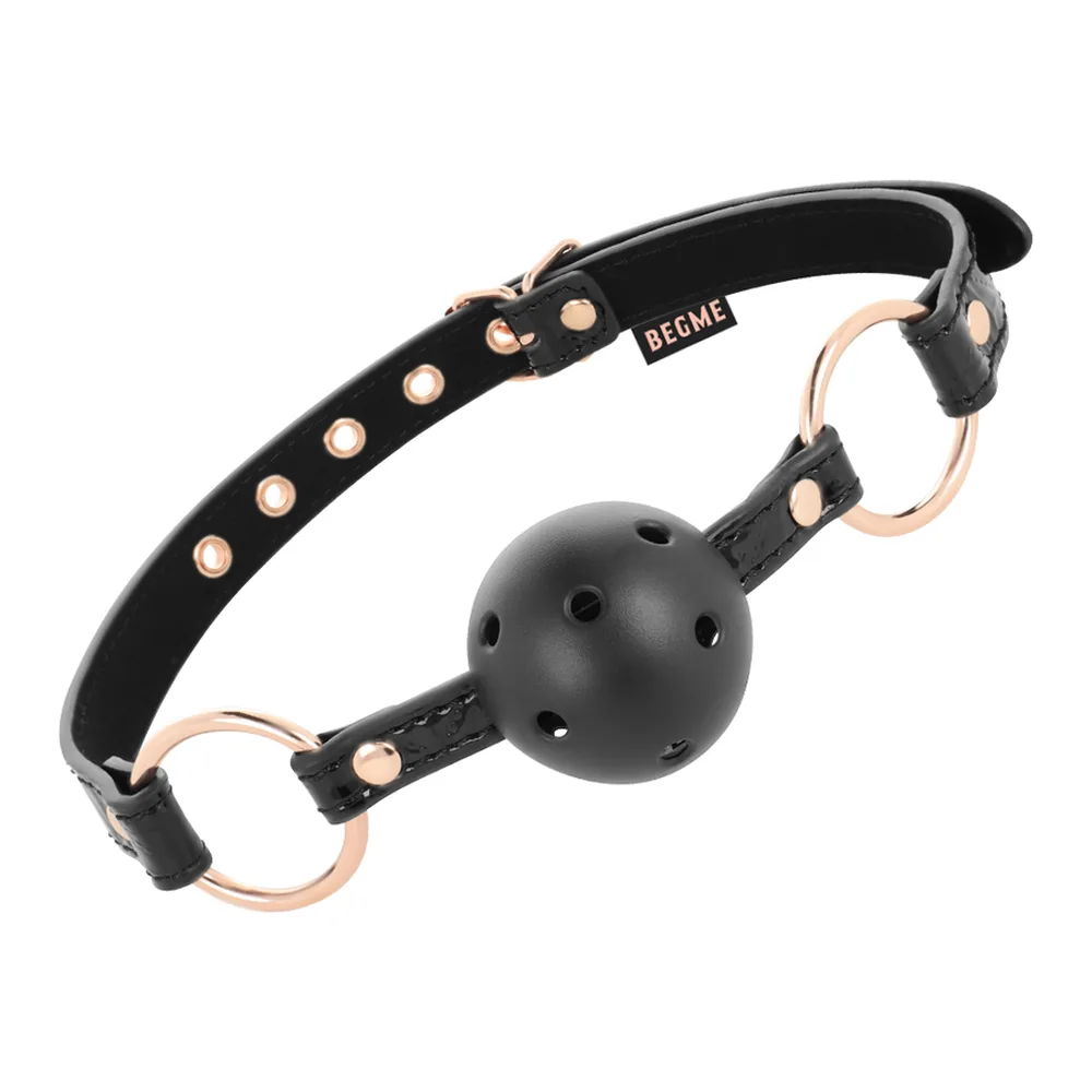 Bâillon boule noir avec sphère ajourée, lanières en similicuir noir ornées de clous dorés et anneaux de métal rose gold, marque BEGME visible