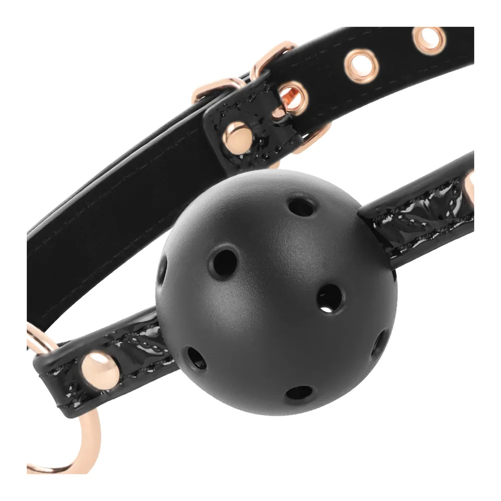 Bâillon boule noir avec sphère ajourée, lanières en similicuir noir ornées de clous dorés et anneaux de métal rose gold, marque BEGME visible