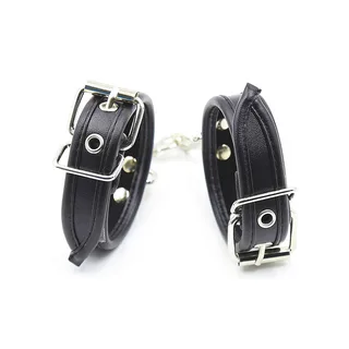 Paire de manchettes de poignets en cuir noir avec boucles métalliques argentées, rivets argentés et mousquetons détachables reliés par une chaîne, sur fond blanc