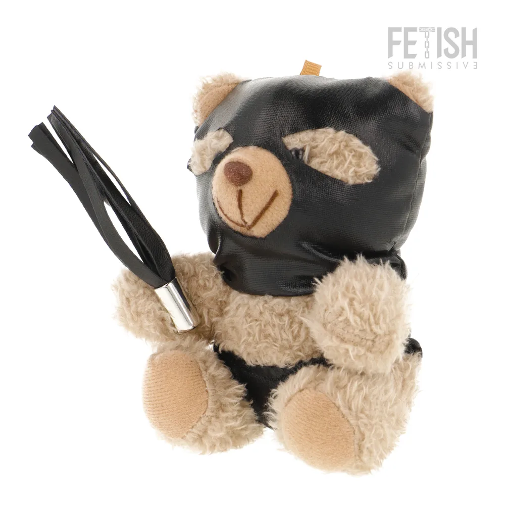 Peluche ours beige et noir avec masque en cuir noir sur le museau, oreilles en dentelle beige, tenant un fouet à lanières noires, sur fond blanc