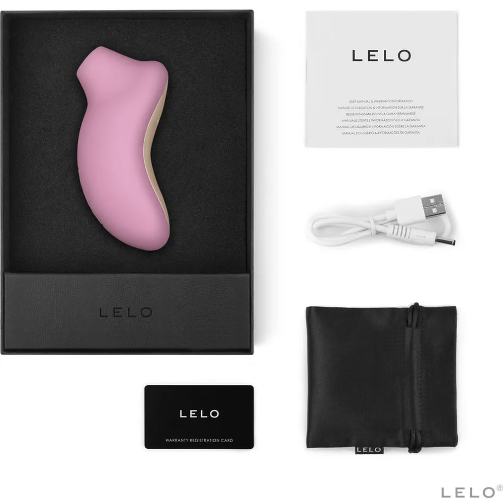 Stimulateur clitoridien rose pâle au design ergonomique bombé, présenté dans son emballage noir avec accessoires : câble USB blanc, pochette de rangement noire et carte de garantie