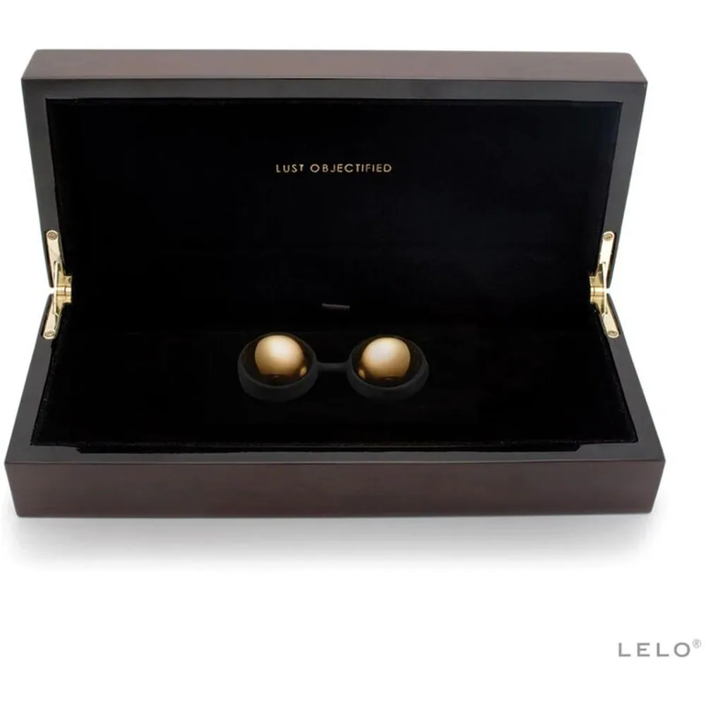 Boîte de présentation gris anthracite ouverte contenant deux boules de Geisha lisses, dorées, nichées dans un écrin noir, avec poignées dorées et inscription "LUST OBJECTIFIED"