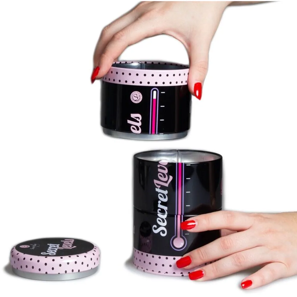 Jeu des Niveaux Secrets avec trois boîtes cylindriques noires à couvercle rose à pois, empilables, accompagnées de jetons de couleur rose et gris. Les boîtes portent des inscriptions et symboles, présentées à la main et groupées sur fond blanc