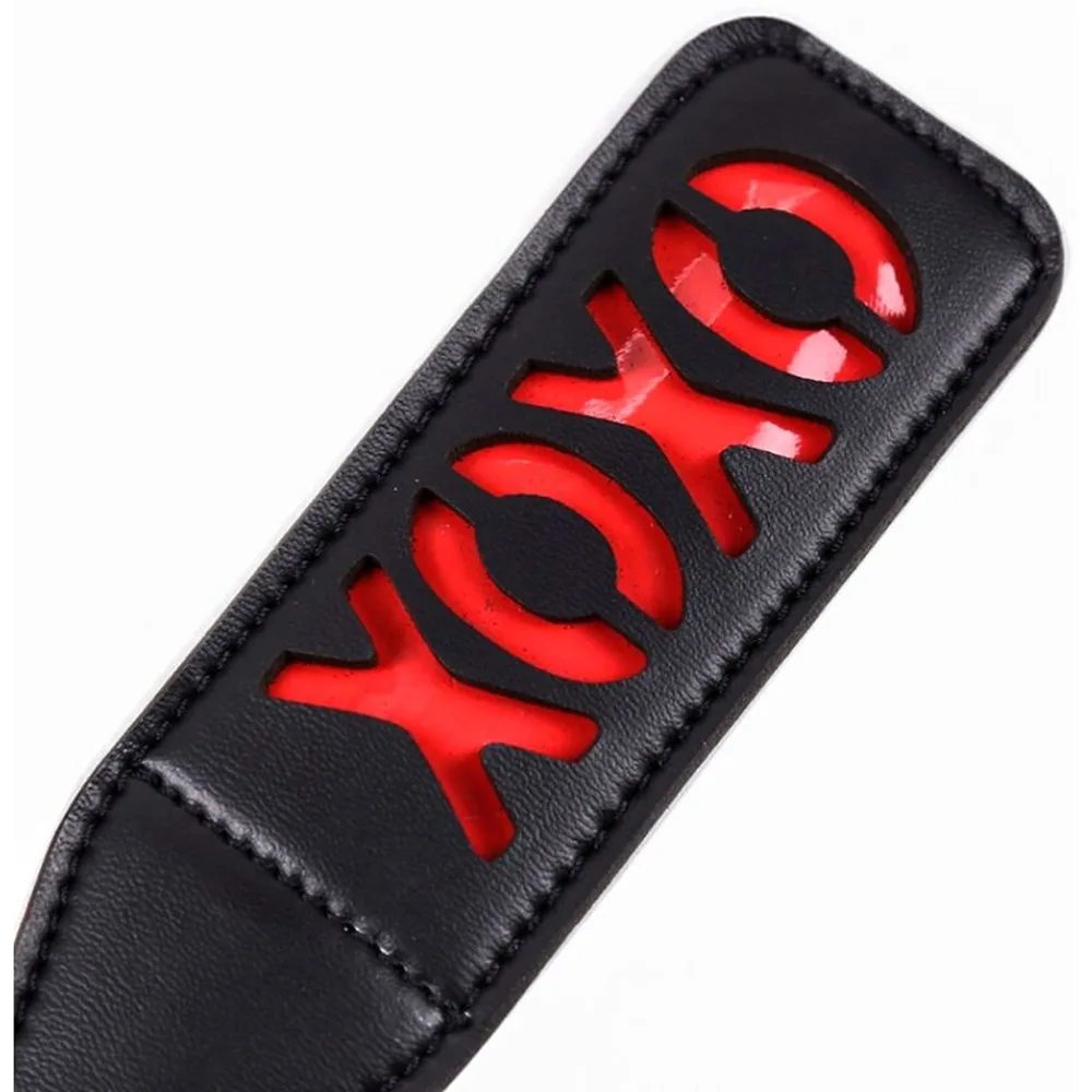 Fouet paddle noir en cuir avec lame rectangulaire ajourée aux lettres XOXO en rouge, manche ergonomique et lanière de suspension noire